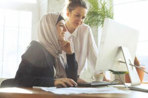 beautiful Arabian woman in hijab working at large computer monitor with colleague standing behind